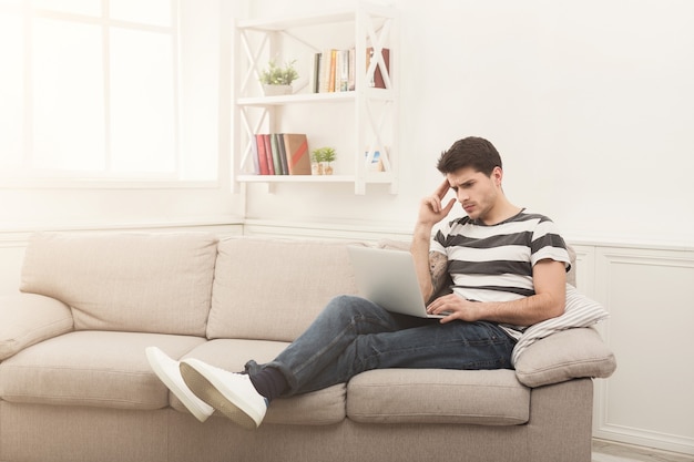 Young serious man at home chatting online on laptop. Thoughtful guy writing article, sitting on beige couch, freelance and remote work concept, copy space