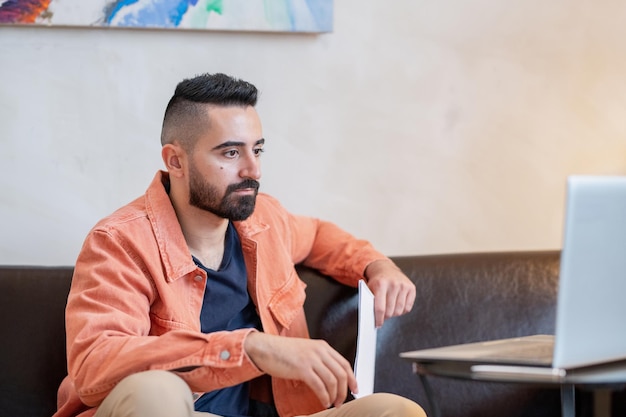 Young serious man looking at laptop screen while sitting on sofa in home environment during online lesson