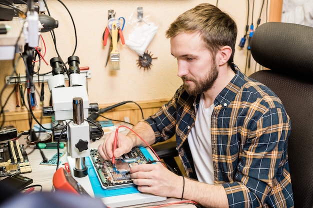 Giovane meccanico serio con due saldatori che riparano il touchpad smontato dal posto di lavoro in officina