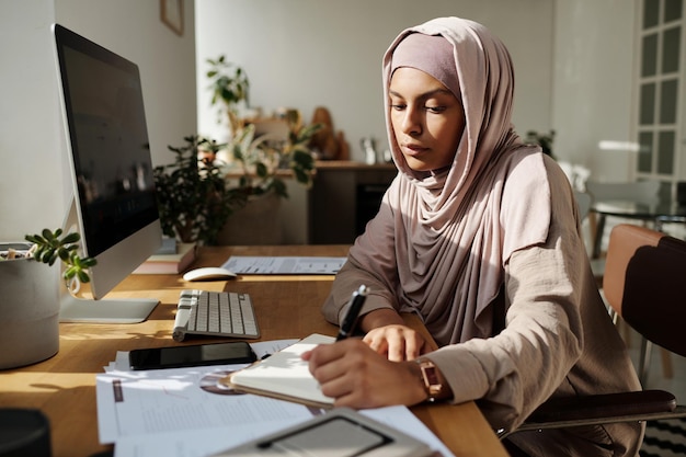 Young serious Muslim businesswoman making notes in notepad ordocument