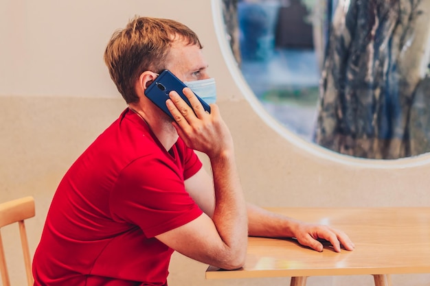 Young serious pensive attentive mean in medical face mask tense thinking speak answer phone in red tshirt in cafe Real people expression life problems solving communication during Covid Pandemic