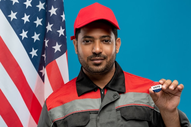 Young serious repairman of Hispanic ethnicity holding vote insignia against stars-and-stripes flag