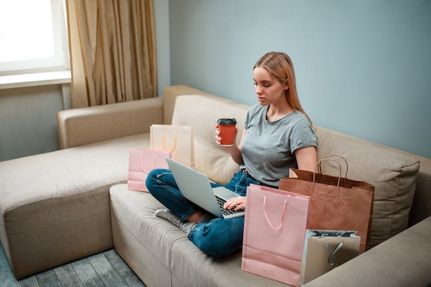 Young serious shopper with coffee is using laptop while sitting on sofa with shopping bags