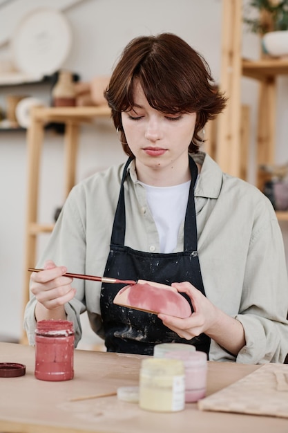 Young serious skilled female artisan painting handmade earthenware