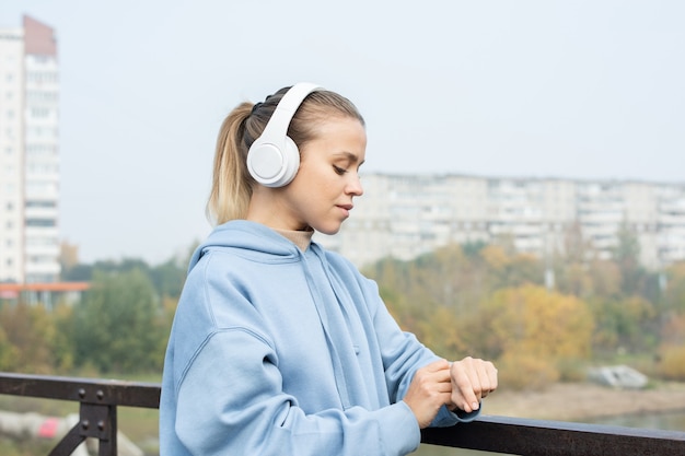 Young serious sportswoman in headphones and hoodie looking at wristwatch while standing by metallic fence in front of camera