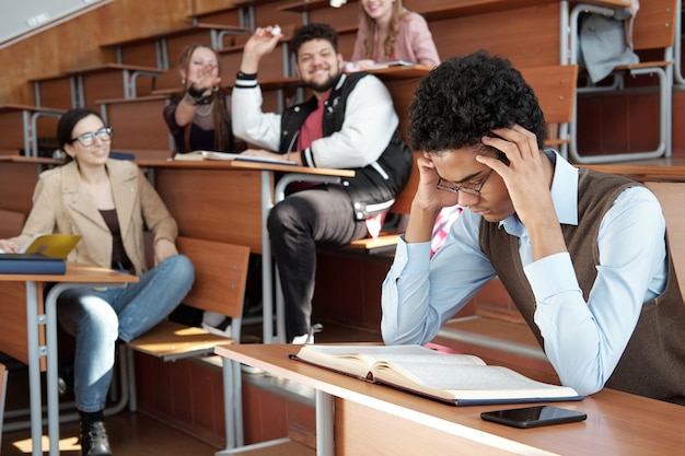 Giovane studente serio in abbigliamento casual chinandosi sul libro aperto sulla scrivania mentre si concentra sulla lettura del passaggio e sull'apprendimento a memoria