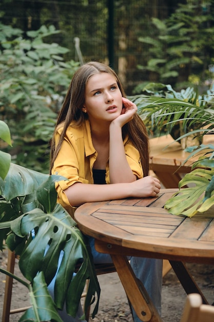 Young serious teenage girl in yellow shirt leaning head on hand thoughtfully looking up while sitting around green leaves in city park