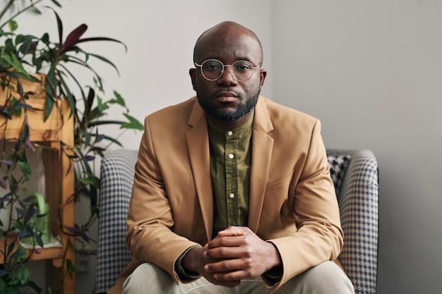 Young serious welldressed male psychotherapist sitting in armchair in office