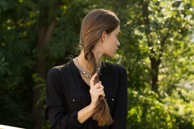 Young serious woman fixing her long brown hair on the background of green trees