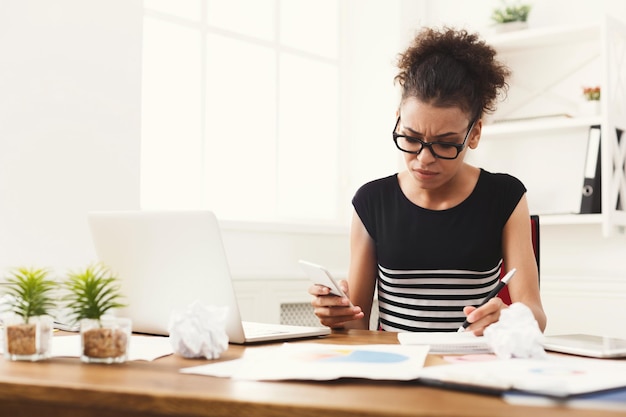 Young serious woman making notes and using smartphone at modern office copy space.