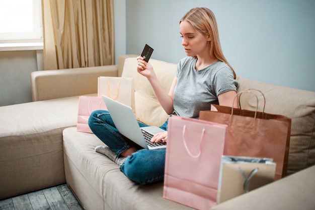 Young serious woman with credit card is looking for big sales in online shop while sitting on a sofa with shopping bags