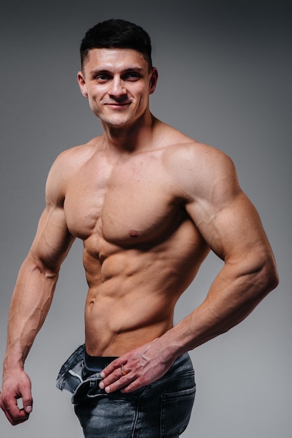 A young sexy athlete with perfect abs poses in the studio topless in jeans on the background. Healthy lifestyle, proper nutrition, training programs and nutrition for weight loss.