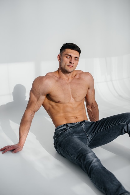 Un giovane atleta sexy con addominali perfetti si siede sul pavimento in studio in topless in jeans sullo sfondo. stile di vita sano, corretta alimentazione, programmi di allenamento e alimentazione per dimagrire.