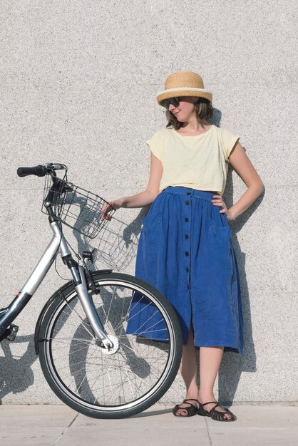 若いセクシーなブロンドの女の子が青いスカートと帽子で自転車の近くに立っています
