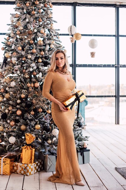 Young sexy blonde in a long gold dress holding a gift near the christmas tree