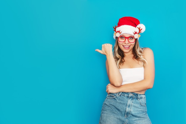 A young sexy blonde woman in a Christmas Santa hat points at empty copy space for text or design