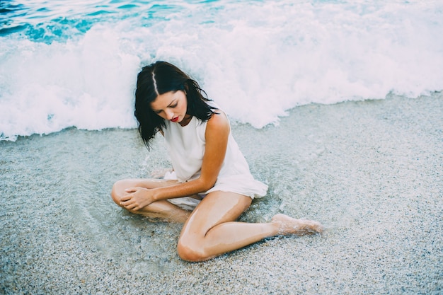 Young sexy brunette in blue water in a white dress on the beach in Greece