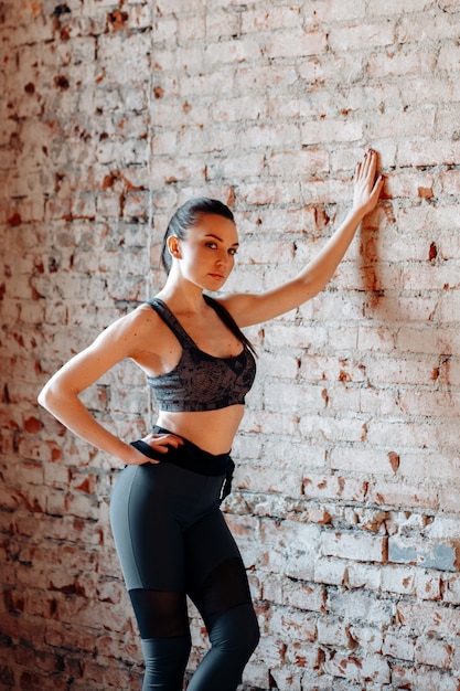 Young sexy brunette woman in sports leggings and top posing against a brick wall in the Studio with loft style.