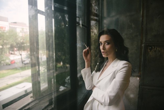 Young sexy brunette woman in suit smoking a cigarette by the window indoors