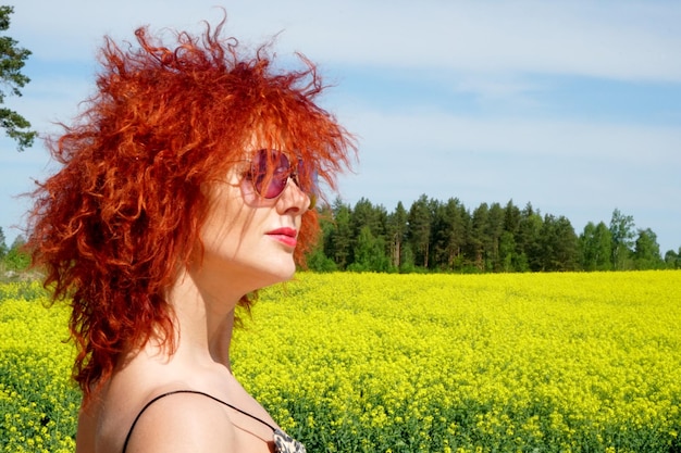 黄色の菜の花畑の背景、夏の日に赤い髪とサングラスの若いセクシーな白人女性