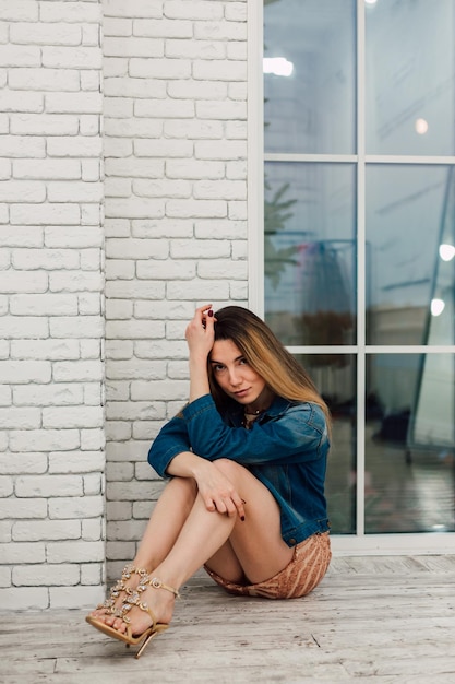 Young sexy girl in a short dress and a denim jacket sits near the panoramic window