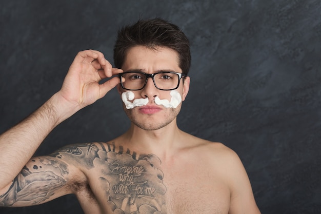 Young sexy tattooed man with moustaches from foam. Topless guy in glasses having fun while shaving, grimacing at camera, gray studio background