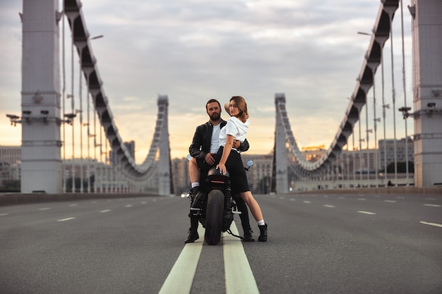Young sexy woman hugging cute man in stylish black leather jacket, sitting on sports motorcycle on the bridge in the city on sunset