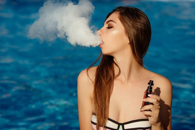 若いセクシーな女性はプールでvapingです。蒸気の雲。ホットでセクシーな女性が水でvaping（電子タバコを吸う）。拡大図。 Vapingのコンセプト