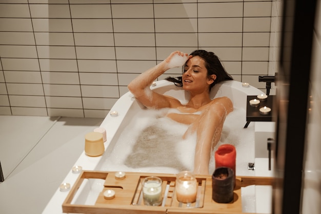 Young and sexy woman relaxing in the cosy bath full of foam and decorated with candles. Healthy treatment and enjoyment