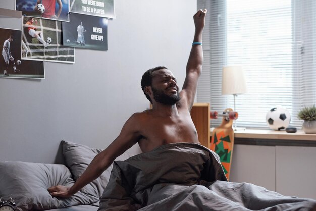 Young shirtless African man stretching in bed after awake