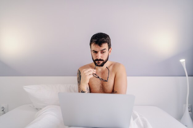 Young shirtless bearded tattooed man sitting on bed in bedroom and using laptop