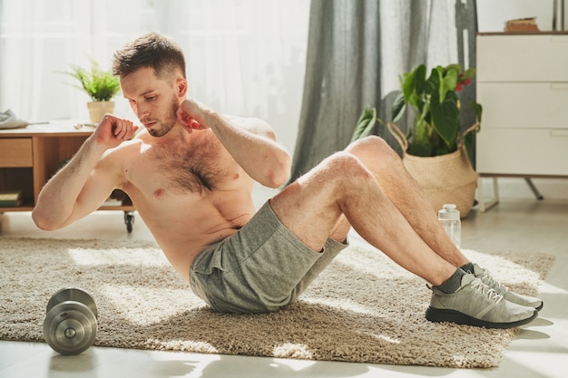 Young shirtless man sitting on carpet in living-room and doing side turns with his legs bent in knee while staying home for quarantine period