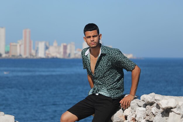 young shirtless man stands behind the sea and wears white pants He is a young Latino with laughing