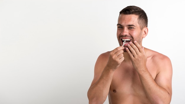 Foto giovane uomo senza camicia che usando filo per i denti per pulire i suoi denti che stanno contro il fondo bianco