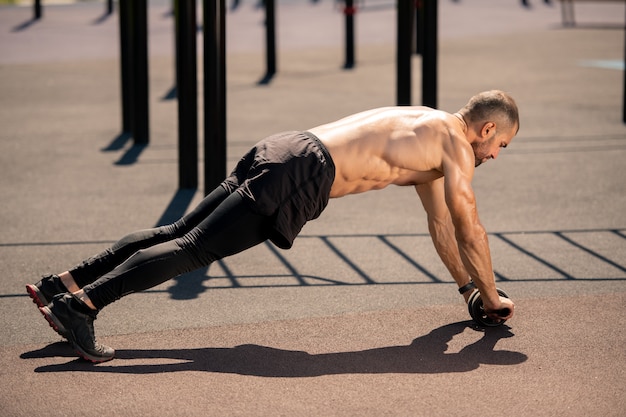 Young shirtless muscular athlete in black shorts, leggins and sneakers bending over sportsground during exercise with rolling trimmer