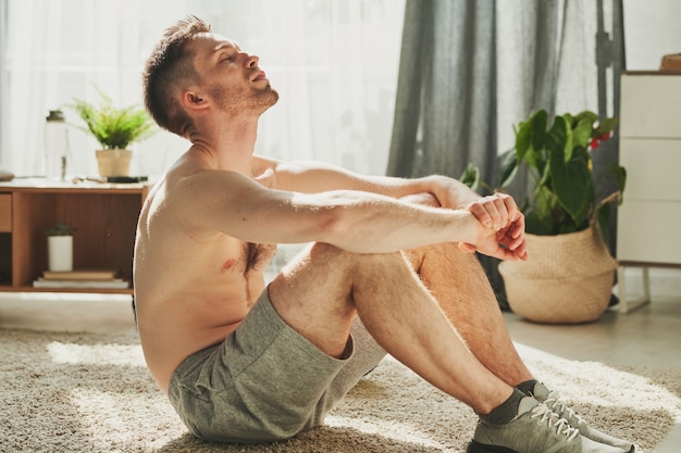 Young shirtless sportsman in sports shoes and shorts sitting on the carpet with his eyes closed and relaxing after hard training at home