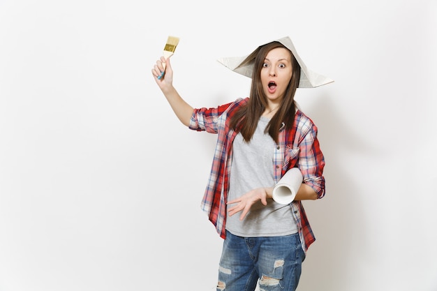 Young shocked beautiful woman in casual clothes and newspaper hat holding paint brush and wallpaper roll isolated on white background. Instruments for renovation apartment room. Repair home concept.