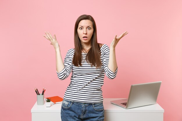 Photo young shocked bewildered woman spreading hands work and standing near white desk with pc laptop isolated on pastel pink background. achievement business career concept. copy space for advertisement.