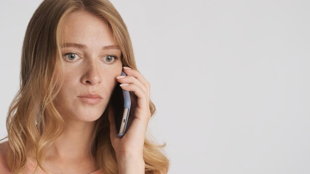 Young shocked blond woman talking on smartphone isolated on white background Astonished expression