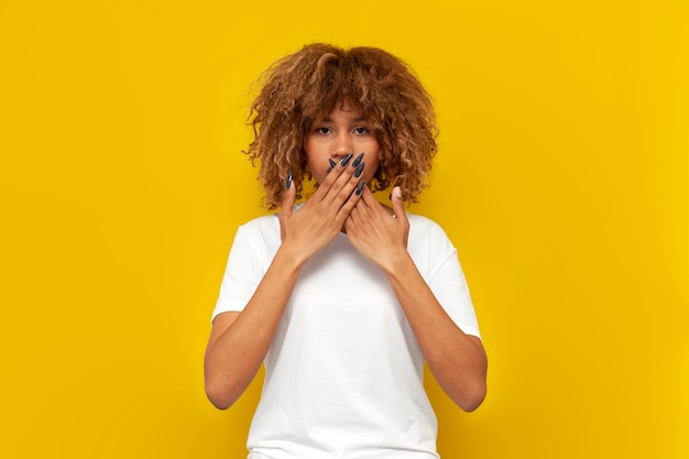 Young shocked curly american girl covers her mouth with her hands on yellow isolated background