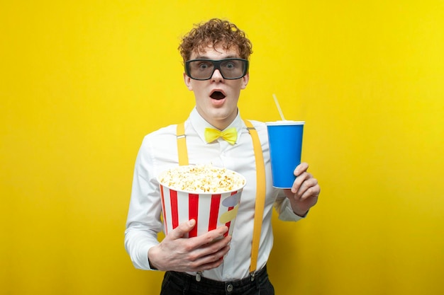 Young shocked guy in festive outfit in 3d glasses watches movie with popcorn and drinks soda