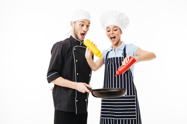 Chef giovane uomo scioccato in uniforme nera tenendo la padella nelle mani mentre la donna cucina in grembiule a strisce utilizzando bottiglie di ketchup e senape su di esso