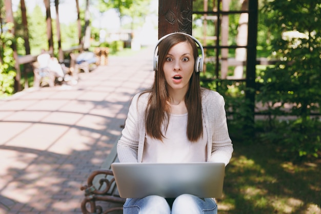 Giovane imprenditrice piuttosto scioccata in abiti casual leggeri. donna che lavora su un moderno computer portatile, ascolta musica in cuffia sulla testa in strada all'aperto. ufficio mobile. concetto di business freelance