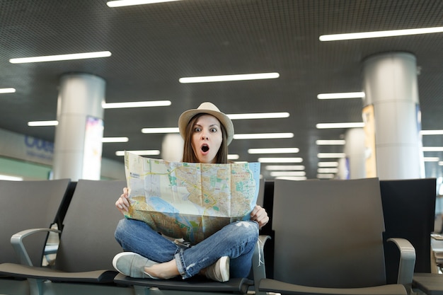 Photo young shocked traveler tourist woman with crossed legs hold paper map, searching route waiting in lobby hall at international airport
