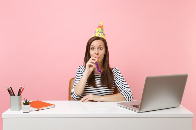 Giovane donna scioccata nel cappello della festa di compleanno con la pipa che suona mentre celebra mentre è seduta a lavorare alla scrivania bianca con un computer portatile