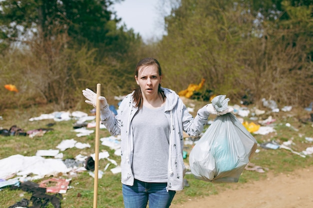 Young shocked woman in casual clothes and latex gloves for cleaning holding trash bags in littered park