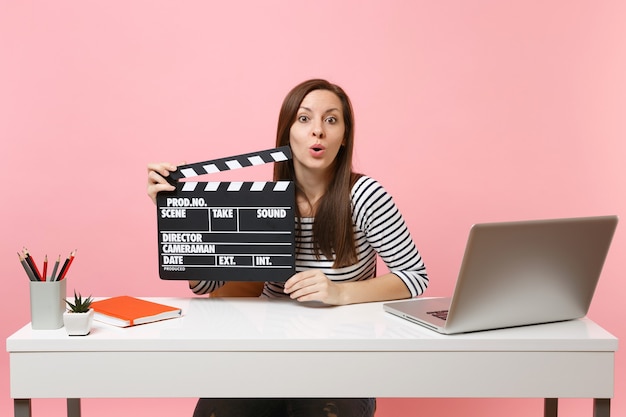 La giovane donna scioccata tiene il classico film nero che fa il ciak lavorando sul progetto mentre si siede in ufficio con il laptop