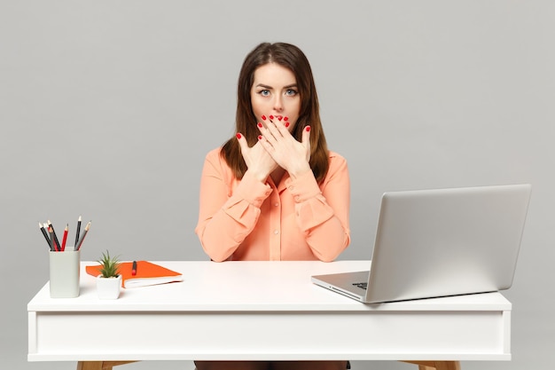 Young shocked woman in pastel casual clothes covering mouth with hands, sit work at desk with pc laptop isolated on gray background. Achievement business career lifestyle concept. Mock up copy space.