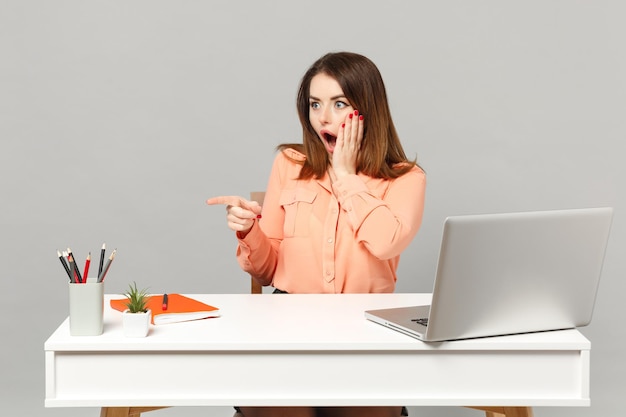 Young shocked woman putting hand on cheek pointing index finger aside, sit work at desk with pc laptop isolated on gray background. Achievement business career lifestyle concept. Mock up copy space.