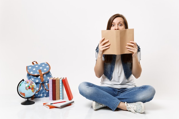 Young shocked woman student in denim clothes covering face with book read sitting near globe, backpack, school books isolated